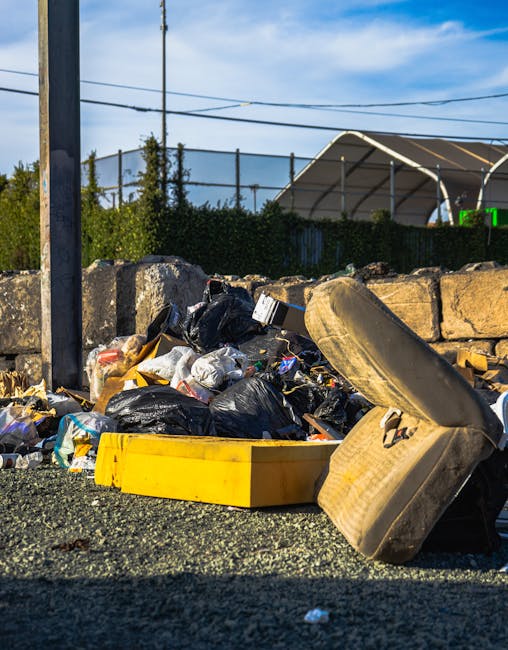 Three large black bin bags filled with waste are placed on a paved sidewalk near a black metal fence. The bags appear to be made of thick plastic and are crumpled with visible creases and some printed